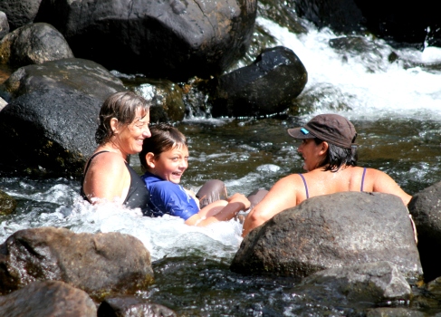 Two women and a child sitting in the flowing water of a river.  Rocks and boulders around them.