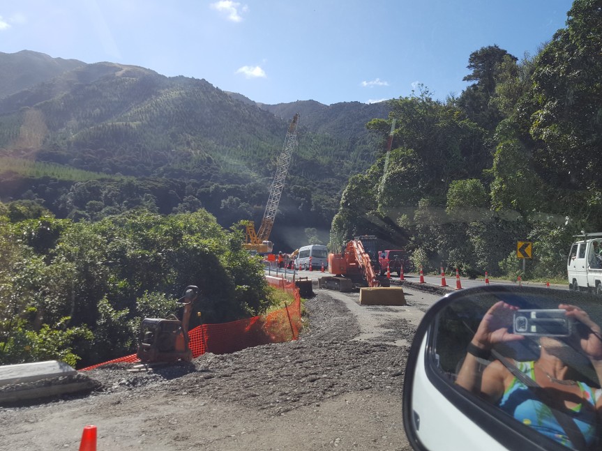 mountain landscape in the background, repairs being made to a road, cranes and diggers working on the road. Bottom right hand corner is the reflection in the wing mirror of the car of the photographer taking the photo.