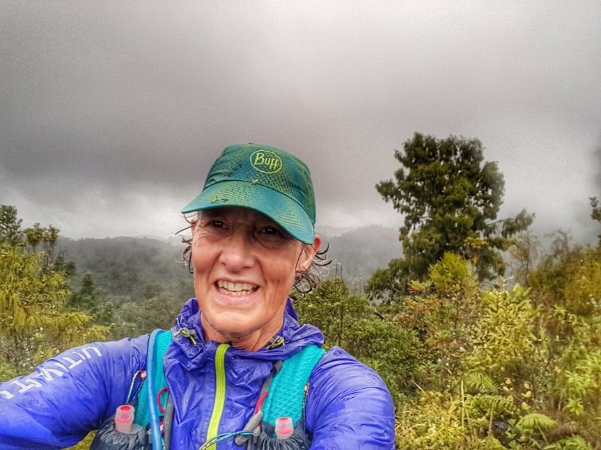 female trailrunner, wearing turquoise cap and purple rain jacket. Self portrait at the top of a hill in the rain with dark clouds and forested valley in the background